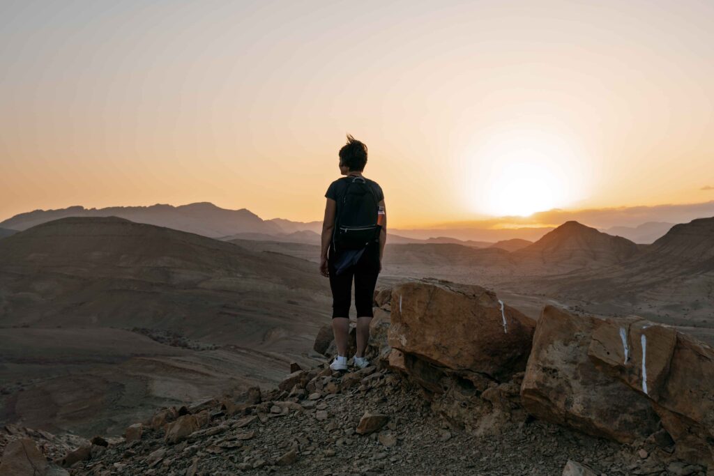 Woman wearing backpack stands alone on the high rock in the dark during sunset and looking at setting sun in the desert mountains of Negev desert in Israel