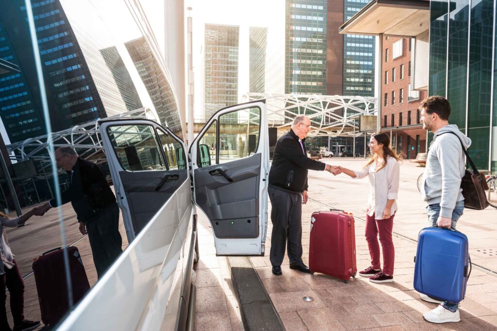 Driver of a airport shuttle minivan, greeting his passengers with their luggage on the sidewalk of a modern city business district