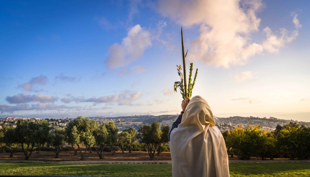 Jewish man in a Tallit praying while waving the Four Species, with a view towards the Temple Mount, the Old City and the Mount of Olives during Sukkot in Israel